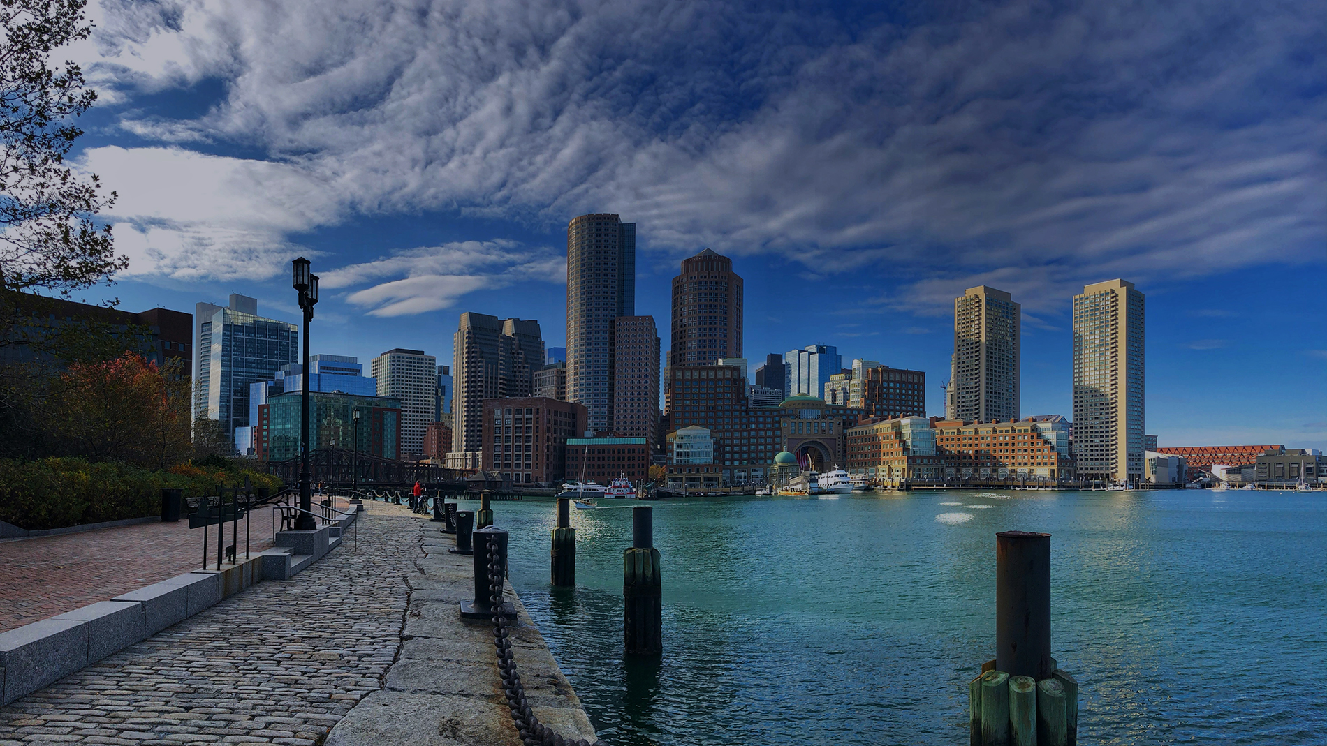 The image shows a view of the Boston skyline from the North End. The sky is blue with some puffy white clouds and the sun is shining brightly. The water in the foreground is calm and there are a few boats docked. On the left side of the image, there is a brick walkway lined with trees and buildings in the background.