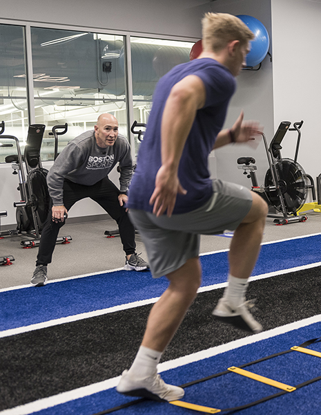 A young male athlete is running through a ladder drill while being watched by his coach. The athlete is wearing a blue shirt and gray shorts, while the coach is wearing a black shirt and gray pants. They are both in a gym.