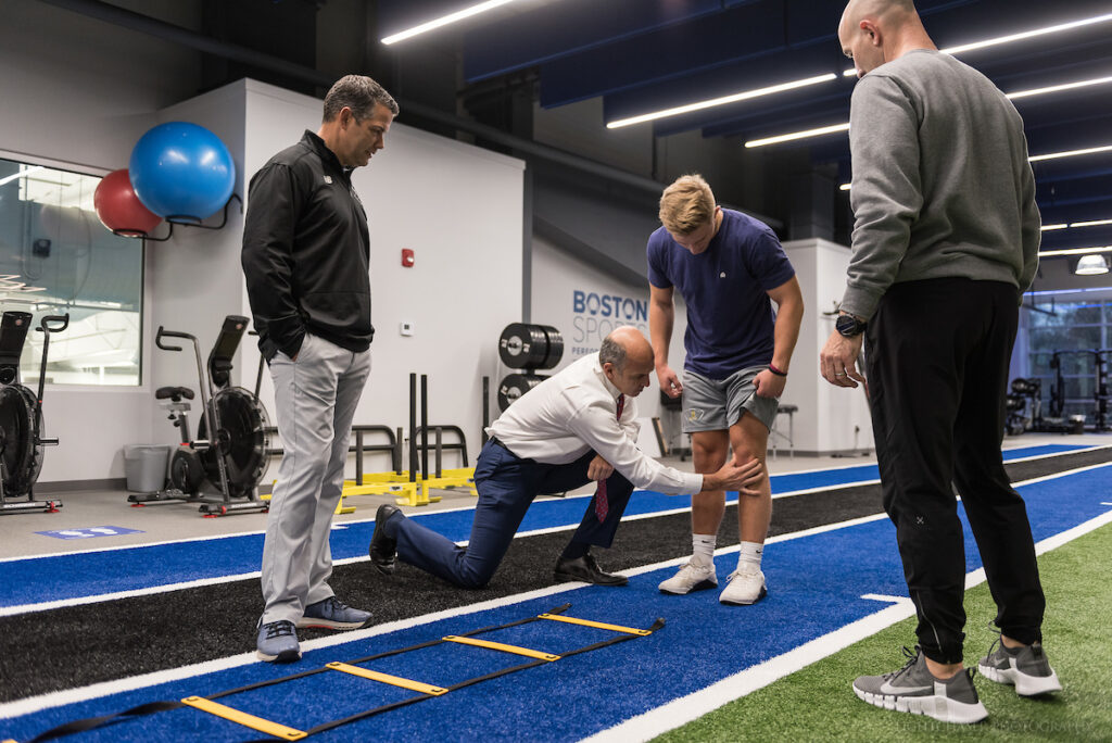 Dr. Gill kneeling down to examine an athlete's knee, while two men are watching them.