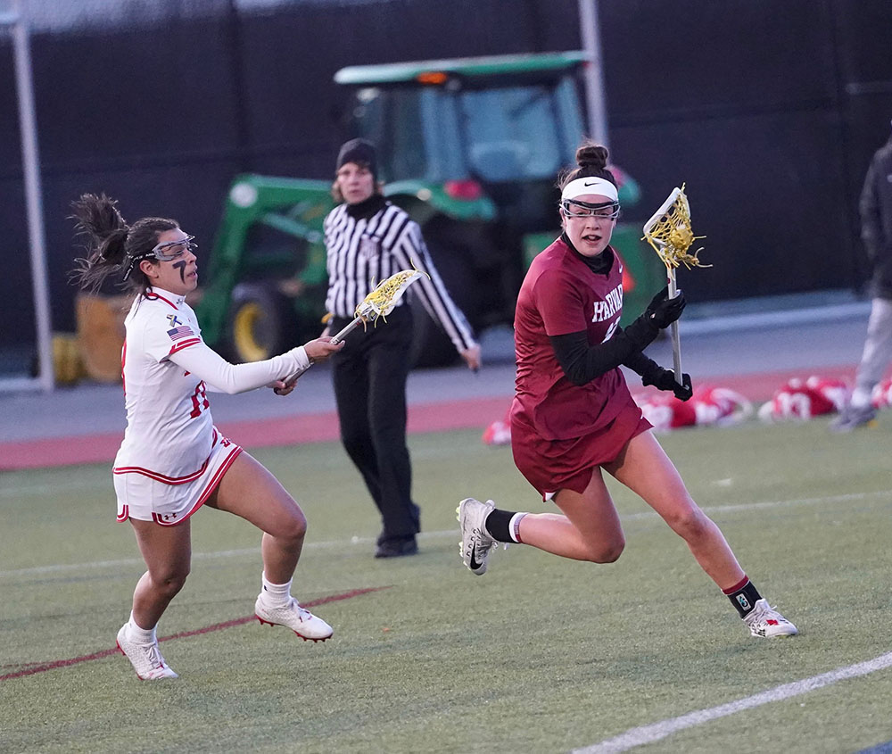 A girl in a red uniform is running with a lacrosse stick in her hand. She is being chased by a girl in a white uniform.